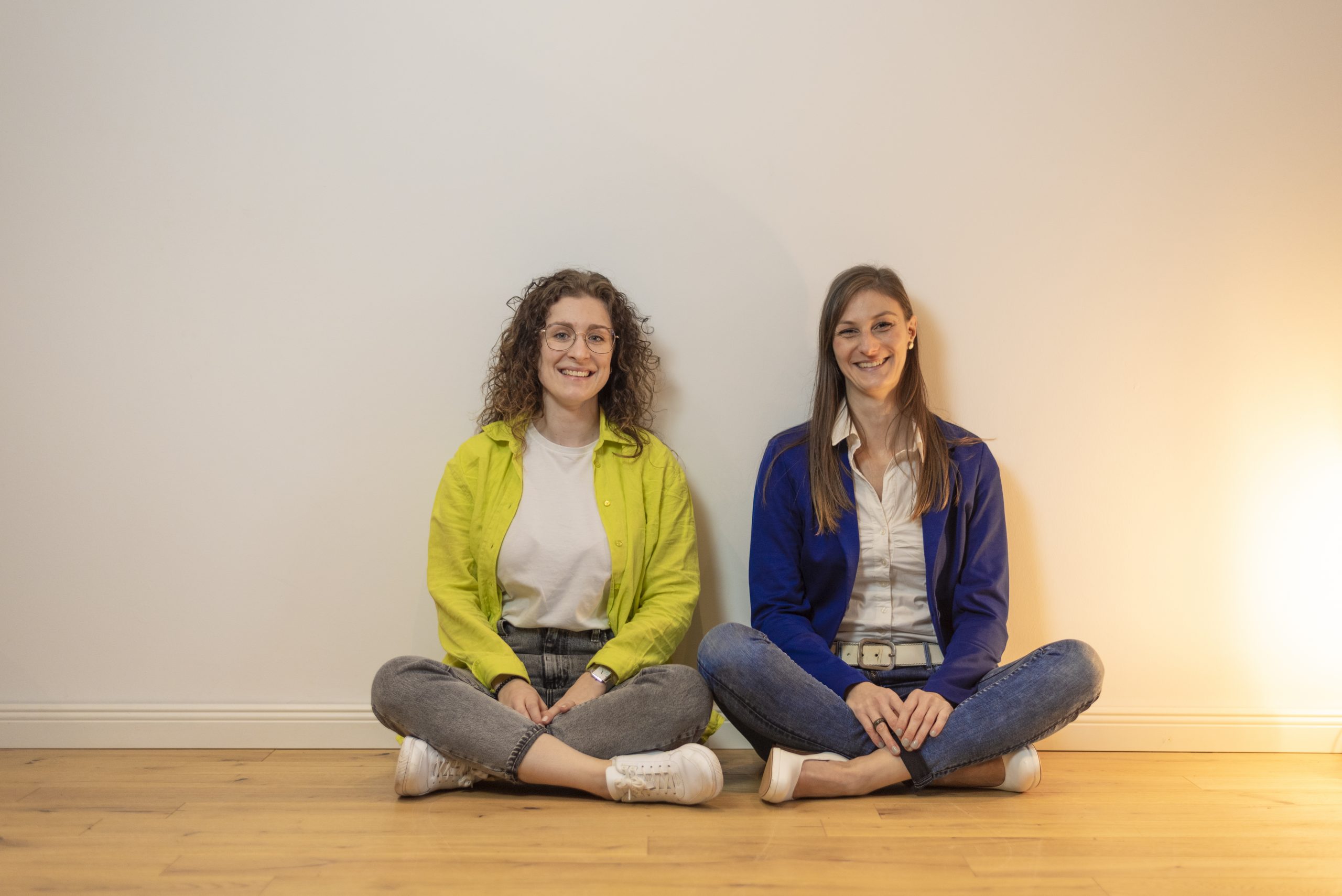 Zwei Frauen sitzen auf dem Boden vor einer beigen Wand.